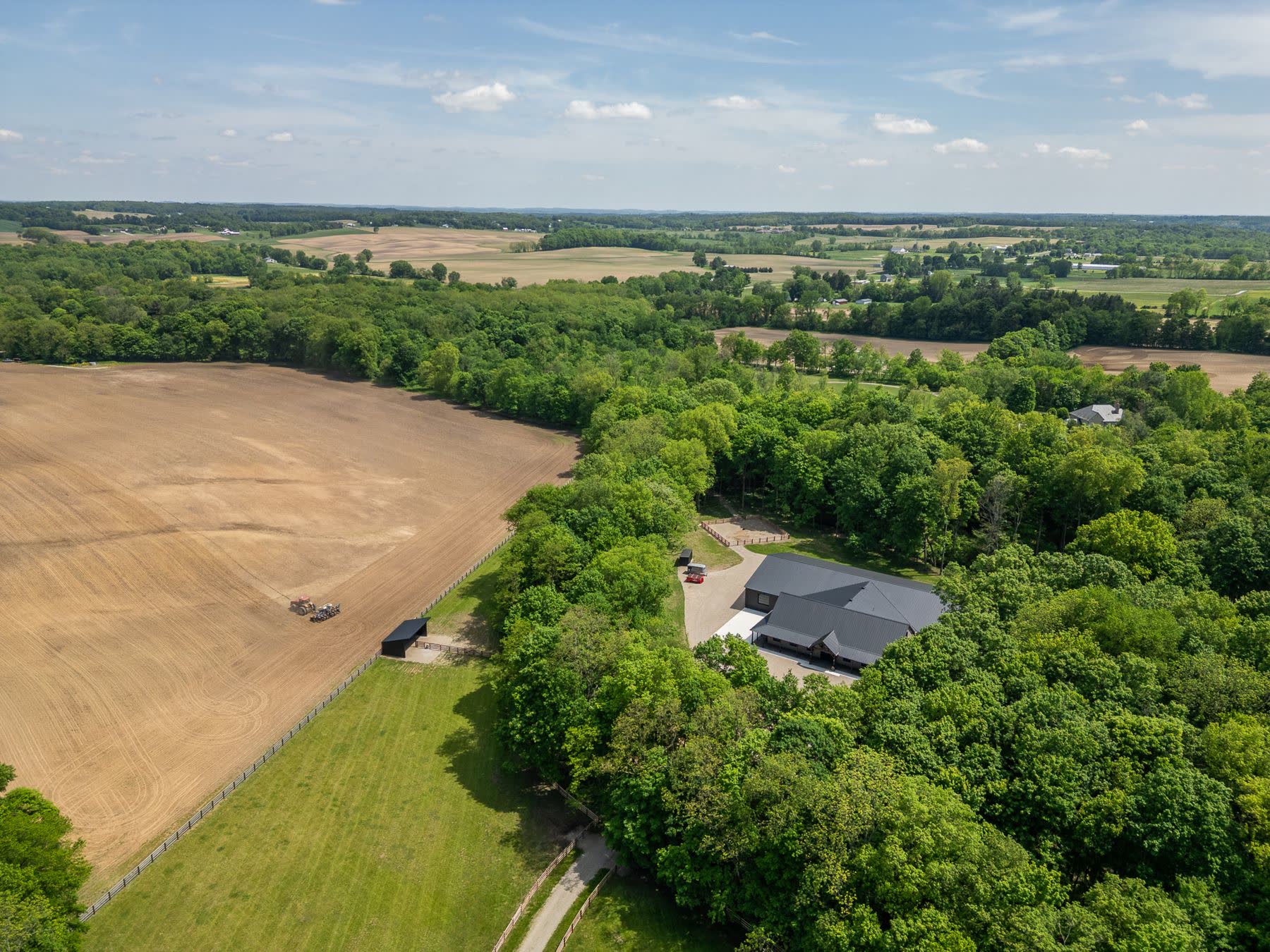 Aerial view of a dark-roofed barn among trees beside open farmland